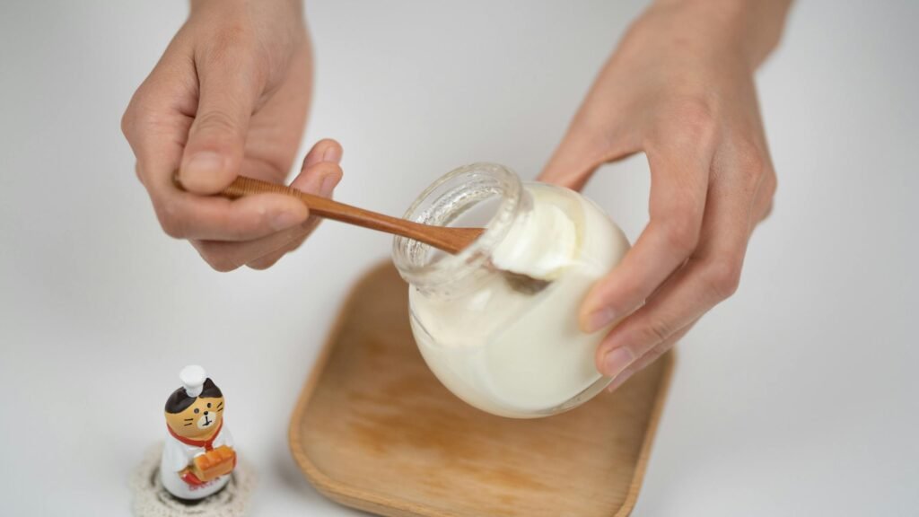 Close-up of hands serving creamy yogurt from a jar using a wooden spoon, with a cute figurine on the side.